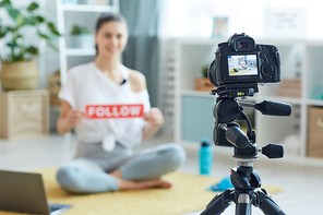 Blurred portrait of smiling young woman holding FOLLOW sign while recording workout video at home and , focus on foreground, copy space