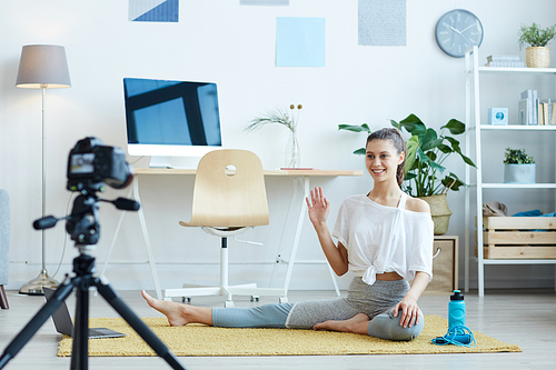 Full length portrait of smiling young woman recording workout video at home and waving at camera, copy space