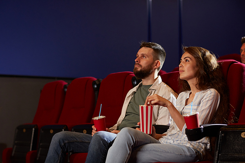 Portrait of modern young couple in cinema watching movie and enjoying popcorn while sitting on red velvet chairs in dark room, focus on smiling woman in foreground, copy space