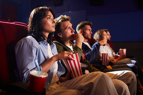 Side view at group of friends watching movie in cinema theater and eating popcorn while sitting in row on red seats, copy space