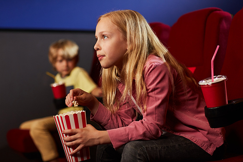 Side view portrait of blonde teenage girl watching movie in cinema theater and eating popcorn, copy space
