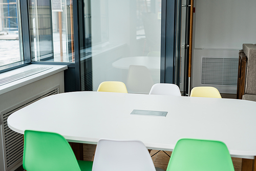 White desk with group of colorful chairs around standing by window of classroom at school