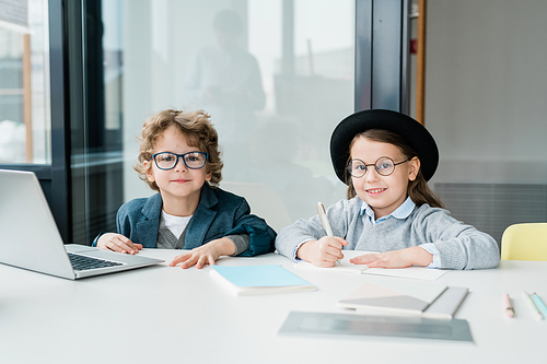 Two youthful diligent schoolkids in casualwear looking at you while sitting by desk in classroom during lesson