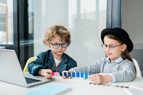 Two friendlyschoolkids in casualwear choosing color from swatch while sitting by desk at lesson
