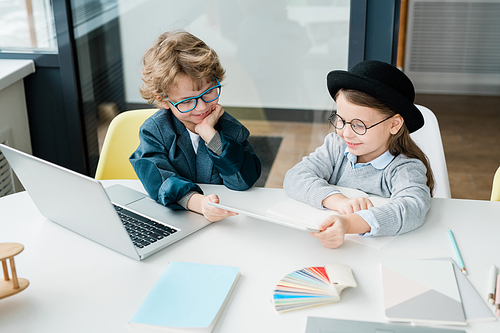 Cute elementary schoolboy and his pretty classmate sitting by desk and watching online video in digital tablet