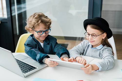 Happy elementary schoolboy pointing at touchpad screen while making presentation to his classmate by desk