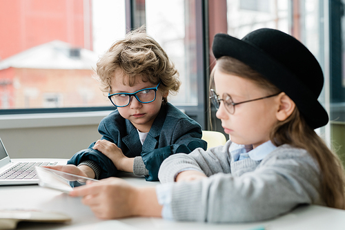Serious schoolboy in eyeglasses looking at his classmate in hat while preparing presentation together by desk