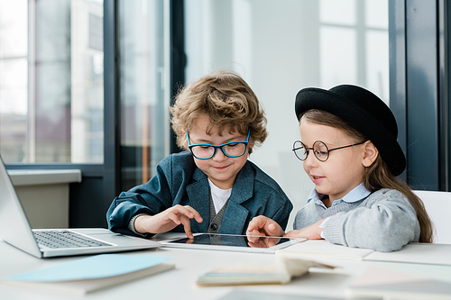 Cute elementary school friends in casualwear and eyeglasses using digital tablet during teamwork at lesson