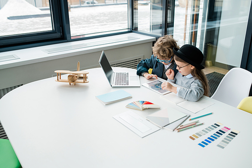 Two cute little designers studying online data in touchpad while working over new creative project by desk