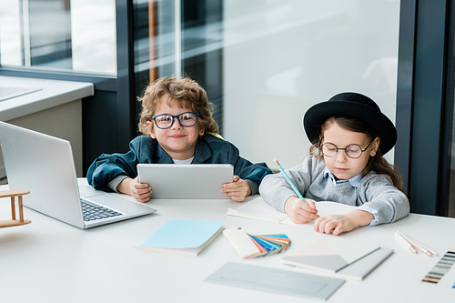 Cute schoolboy with tablet looking at you while sitting by desk next to serious classmate drawing or making notes in copybook