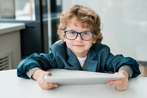 Cute schoolboy with digital tablet looking at you while sitting by desk and preparing homework