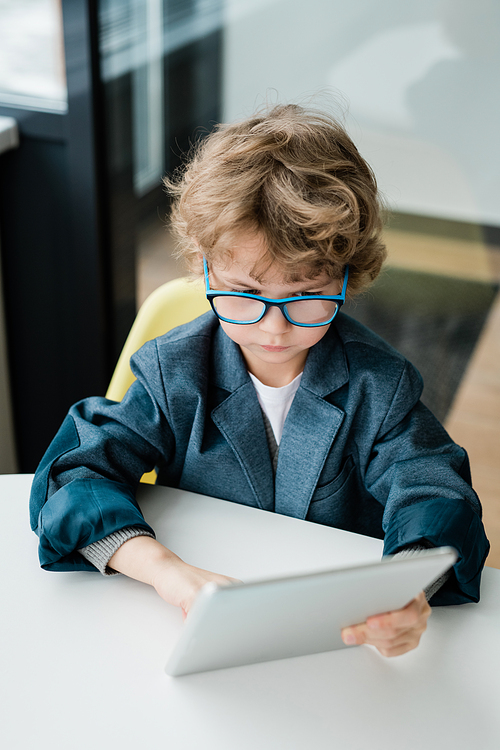 Cute serious schoolboy in eyeglasses looking at display of digital tablet while sitting by desk and preparing homework