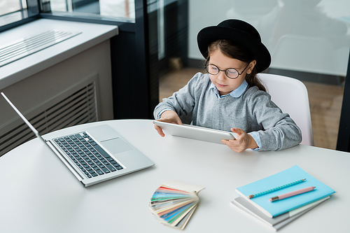 Adorable schoolgirl in eyeglasses looking at display of digital tablet while preparing homework by desk