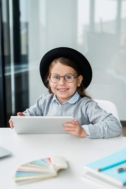 Cute smiling schoolgirl with digital tablet looking at you while sitting by desk and thinking of new ideas