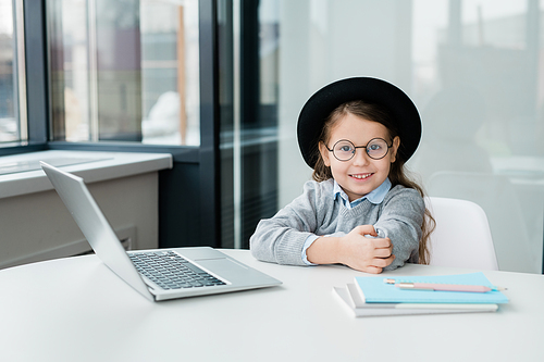 Cute smiling schoolgirl in eyeglasses and casualwear looking at you while preparing homework in front of laptop by desk