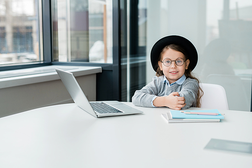 Diligent schoolgirl in eyeglasses and casualwear preparing homework in front of laptop while sitting by desk