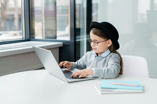 Adorable schoolgirl in casualwear and eyeglasses sitting by desk in front of laptop while preparing homework