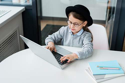 Clever casual schoolgirl in eyeglasses preparing homework while sitting by desk in front of laptop