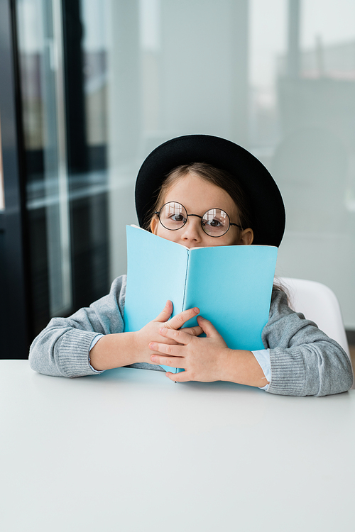 Cute little schoolgirl in eyeglasses and hat looking at you while covering mouth with open book by desk
