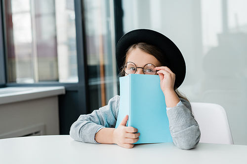 Cute little schoolgirl in eyeglasses and hat peeking out of open book at lesson of literature while sitting by desk