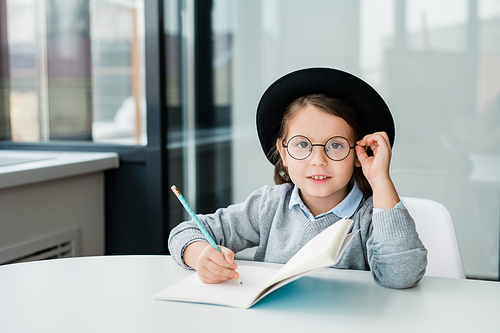 Adorable clever schoolgirl in hat looking through eyeglasses at you while sitting by desk and making notes in copybook