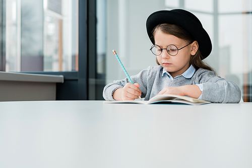 Serious clever schoolgirl in hat sitting by desk and making notes or drawing picture in copybook at lesson