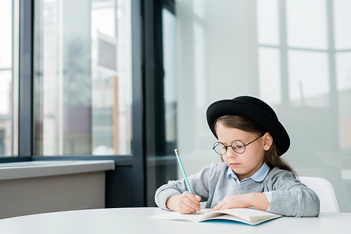 Serious little schoolgirl in hat and eyeglasses making notes or drawing picture in copybook at lesson