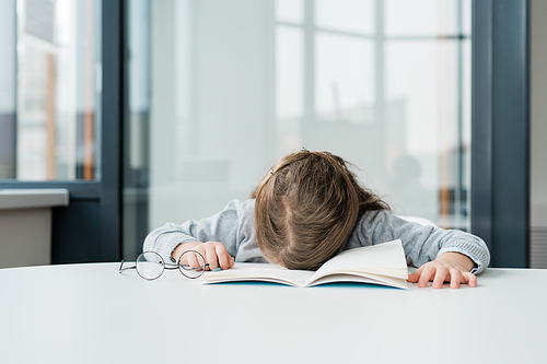 Very tired or bored schoolgirl with eyeglasses napping on open copybook by desk in classroom after lesson