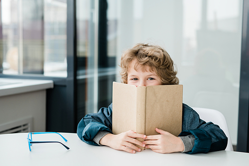 Cute happy little schoolboy peeking out of open book at lesson of literature while sitting by desk