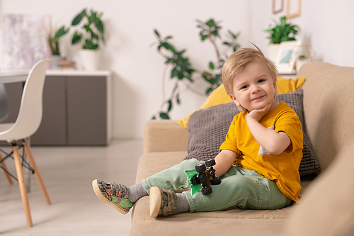 happy cute blond boy in casualwear sitting on soft couch in living-room and playing with toy car in home