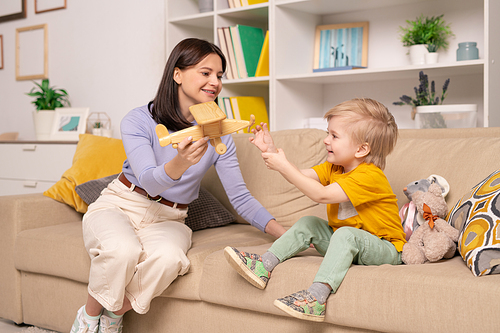 pretty young woman in casualwear showing wooden airplane to her cute little son while playing with him on couch in home