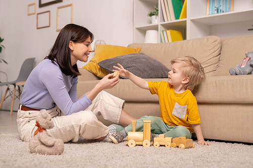 Cute little boy in casualwear sitting on the floor by couch and taking toy cube from hand of his mother while playing with wooden train