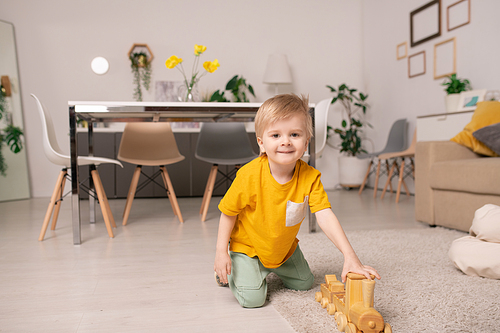 Happy cute little boy playing with wooden train while sitting on rug on the floor on background of table and group of chairs around