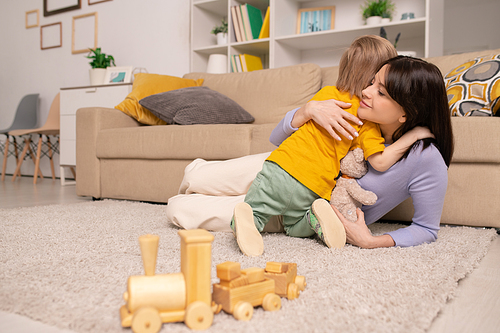 Adorable blond little boy giving hug to his happy mom while both relaxing on the floor by couch at home during period of quarantine