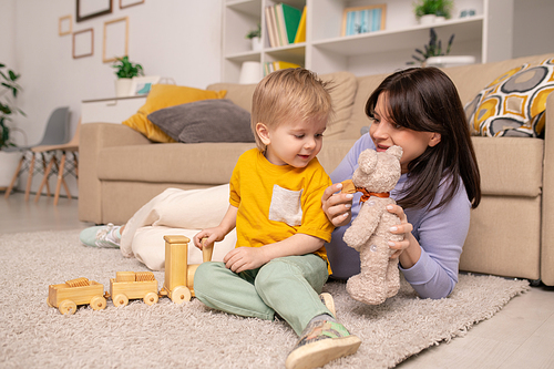 Cheerful young mother lying on carpet and showing toy bear to son while they playing with toys together at home