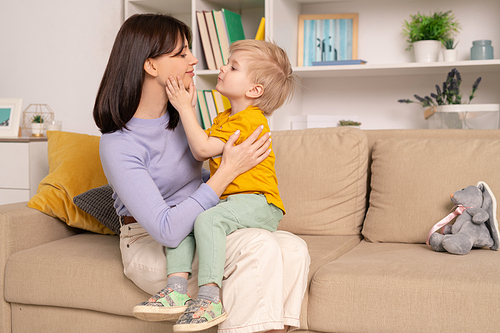 Happy young affectionate mother and her cute little son expressing love to each other while sitting on soft couch in living-room at home