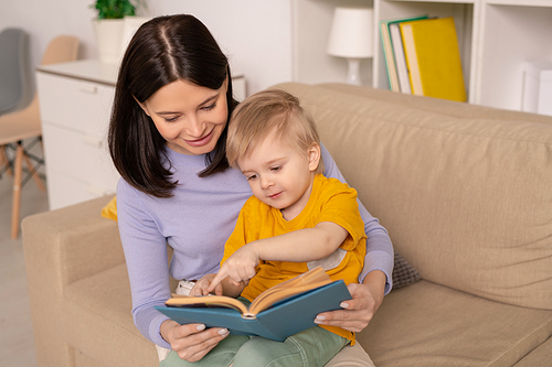 Cute little boy pointing at picture on page of book while discussing one of fairy tales with his mother during period of home isolation