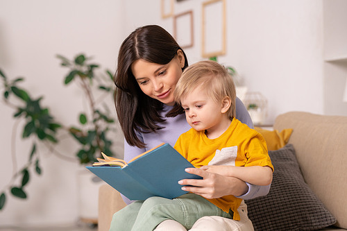 Content beautiful young mother sitting on sofa in living room and reading fairy tale to blond-haired son