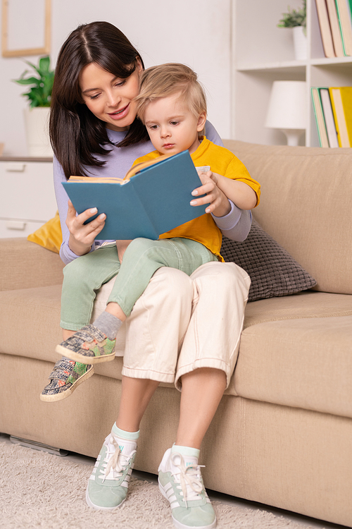 Happy young mother sitting with little son on knees on sofa and reading childrens book with pictures to him