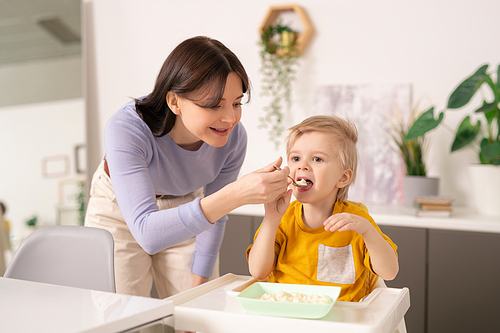 Young careful woman in casualwear bending over small dinner table while helping her cute little son with breakfast in the kitchen