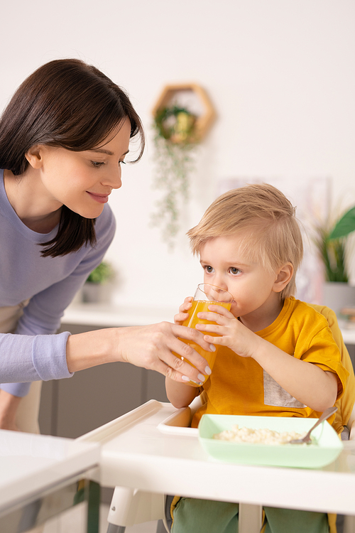 Smiling young mother standing at highchair and giving fresh juice to little son in kitchen