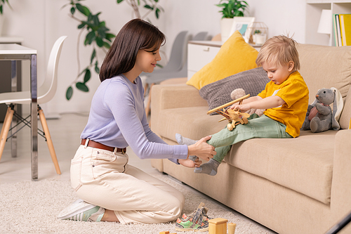 Beautiful young mother in sweater sitting on carpet in living room and putting little son on while he playing with toy plane