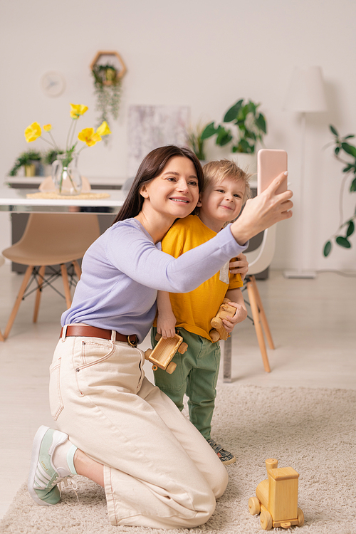 Cheerful young woman and her adorable little son looking at smartphone camera while making selfie at home during self-isolation