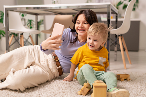 Happy young mother laying on carpet and using smartphone while posing with little son for selfie