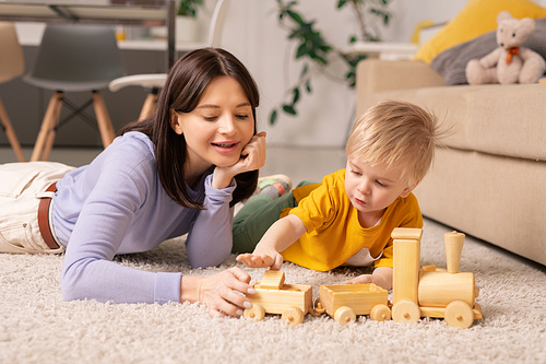 Cute little boy and his young pretty mother lying on fluffy rug on the floor of living-room and playing with wooden train after dinner