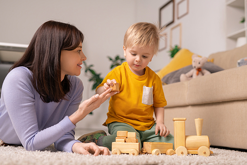 Smiling attractive young mother lying on carpet and giving wooden block to son while teaching him to stack toy blocks