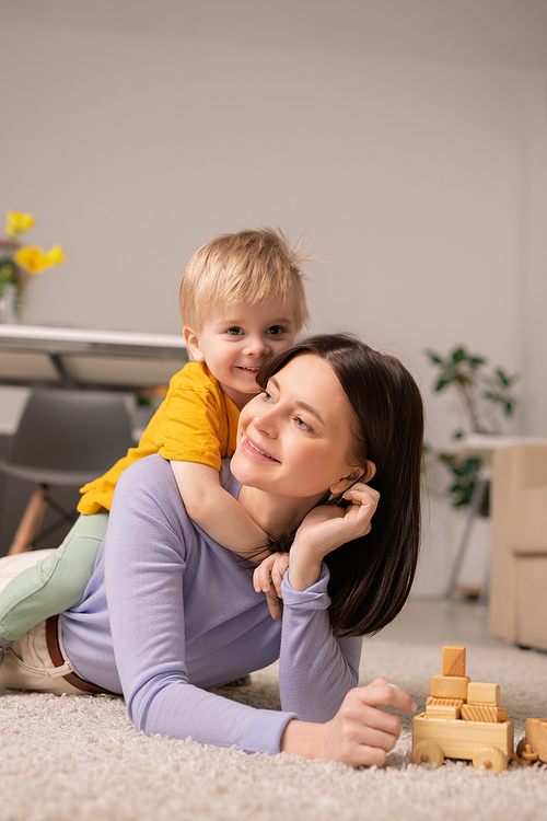 Cute little boy embracing his cheerful mother while both lying on the floor and playing toys together in living-room on quarantine