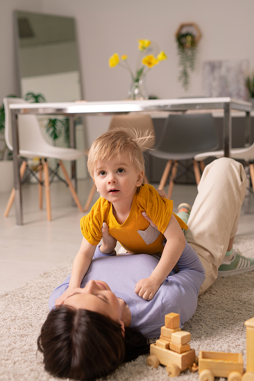 Cute toddler boy lying on mothers chest and looking up while they playing on floor at home