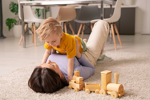 Pretty young brunette woman lying on fluffy rug on the floor and playing with her adorable little son while staying at home during quarantine