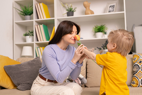 Happy attractive young mother sitting on sofa and smelling flower given by son at home